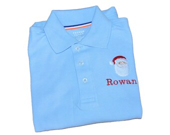 May include: A light blue polo shirt with a collar and three buttons. The shirt features an embroidered Santa Claus face wearing a red hat and the name Rowan stitched in red thread below it. The inner collar tag reads French Toast.