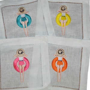 May include: Set of four white linen cocktail napkins with embroidered images of a woman in a swimsuit and a straw hat floating on a colorful inflatable ring. The rings are turquoise, yellow, pink, and orange.