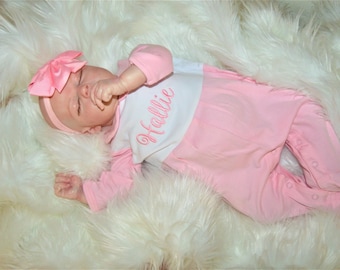 May include: A pink and white baby sleeper with the name "Hallie" embroidered on the front. The sleeper is worn by a baby doll lying on a white fluffy surface.