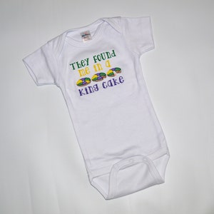 May include: White baby onesie with green, yellow, and purple embroidery that reads "They found me in a king cake".