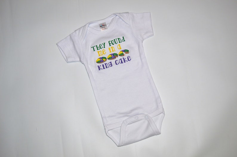May include: White baby onesie with green, yellow, and purple embroidery that reads "They found me in a king cake".