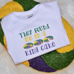 May include: White baby onesie with the text "They found me in a king cake" embroidered in green, purple, and gold. The onesie is on top of a purple, green, and gold king cake.