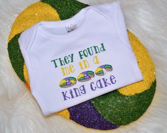 May include: White baby onesie with the text "They found me in a king cake" embroidered in green, purple, and gold. The onesie is on top of a purple, green, and gold king cake.