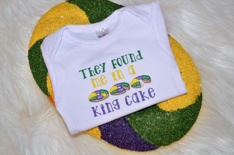 May include: White baby onesie with the text "They found me in a king cake" embroidered in green, purple, and gold. The onesie is on top of a purple, green, and gold king cake.