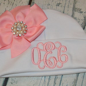May include: White baby hat with a pink bow and a rhinestone embellishment. The hat is embroidered with the monogram "O.C.C."