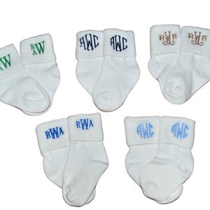 May include: Five pairs of white infant socks, each featuring a unique embroidered monogram on the cuff. The monograms are stitched in various colors including green, dark blue, brown, and light blue, displaying letters such as RWA and AWC.