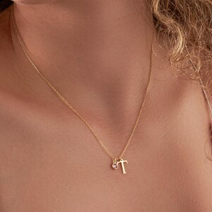 May include: A gold-colored necklace with a delicate chain. The necklace features the letter "T" pendant and a small pink gemstone charm. The necklace is worn around the neck, showcasing its simple elegance and personalized design.