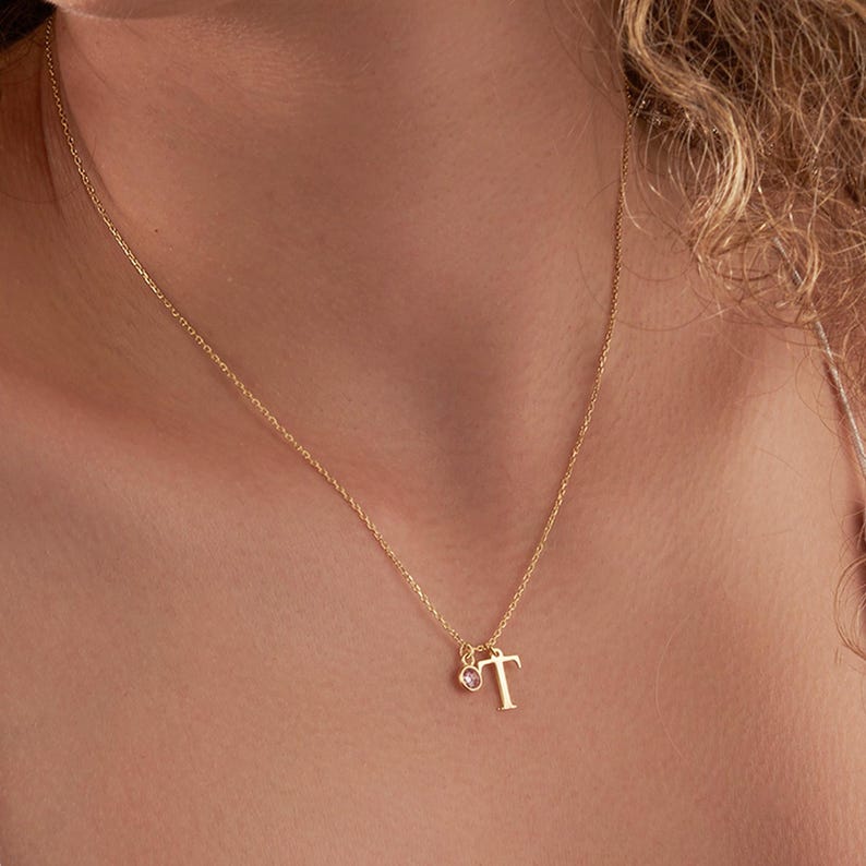 May include: A gold-colored necklace with a delicate chain. The necklace features the letter "T" pendant and a small pink gemstone charm. The necklace is worn around the neck, showcasing its simple elegance and personalized design.
