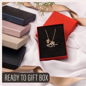 May include: A gold-colored necklace with the initials "BR" and small charms is displayed in a red gift box with a black interior. Several other gift boxes in various colors are stacked nearby. The text "READY TO GIFT BOX" is at the bottom.