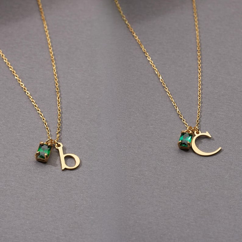 May include: Two gold-colored necklaces with delicate chains. Each necklace features a small, emerald-green rectangular gemstone and a gold letter pendant. One pendant is the letter "b", and the other is the letter "c".