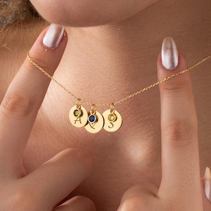 May include: A gold necklace featuring three disc pendants, each with a gemstone and an engraved letter. The gemstones are green, blue, and yellow. The necklace is held up by a person.