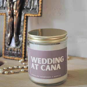 May include: A glass jar candle with a gold lid and a purple label. The label reads "WEDDING AT CANA" with "Sparkling Champagne & Berries" below. The candle sits on a wooden surface.