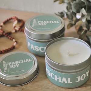 May include: Three round, silver-colored metal tins of candles with a light blue label that reads "PASCHAL JOY" in white text. The candle inside is white. The scent is "Yuzu Blossom". A beaded bracelet is in the background.