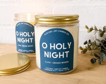 May include: Two clear glass jar candles with gold metal lids. Each candle features a blue arched label with the white text O HOLY NIGHT, PASCHAL JOY CANDLE CO, CLOVE SMOKED WOODS, COCONUT APRICOT CANDLE, HANDMADE IN CT, and 207G / 7.3 OZ.