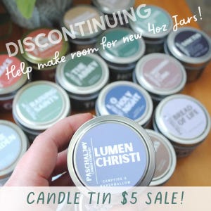 May include: A collection of round metal candle tins with colorful labels featuring various names and scents. The labels include text such as Lumen Christi, Raising Saints, and Bread of Life. The text DISCONTINUING Help make room for new 4oz Jars! is visible.