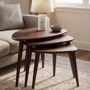 May include: A set of three dark brown, oval-shaped nesting tables with tapered legs. A white marble lamp with a cream shade and a small, round bowl sit on top of the tables. The tables are arranged in front of a light-colored sofa.