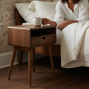 May include: A mid-century modern style walnut nightstand with a drawer and open shelf. The nightstand has tapered legs and a rounded top. A person is sitting on a bed next to the nightstand, holding a mug.