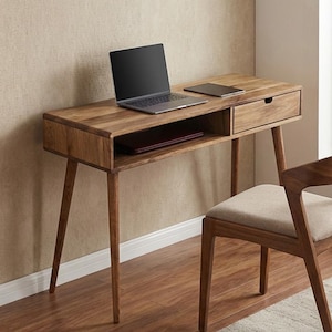 Puede incluir: Un escritorio de madera con un portátil, una tableta y un cajón. Una silla de madera con un cojín beige está al lado del escritorio. El escritorio tiene un estante inferior y es de madera marrón claro.