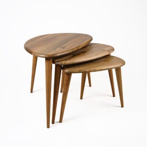 Op de afbeelding: Een set van drie nesttafels gemaakt van hout met een natuurlijke afwerking. De tafels hebben een unieke ovale vorm en worden ondersteund door slanke poten.