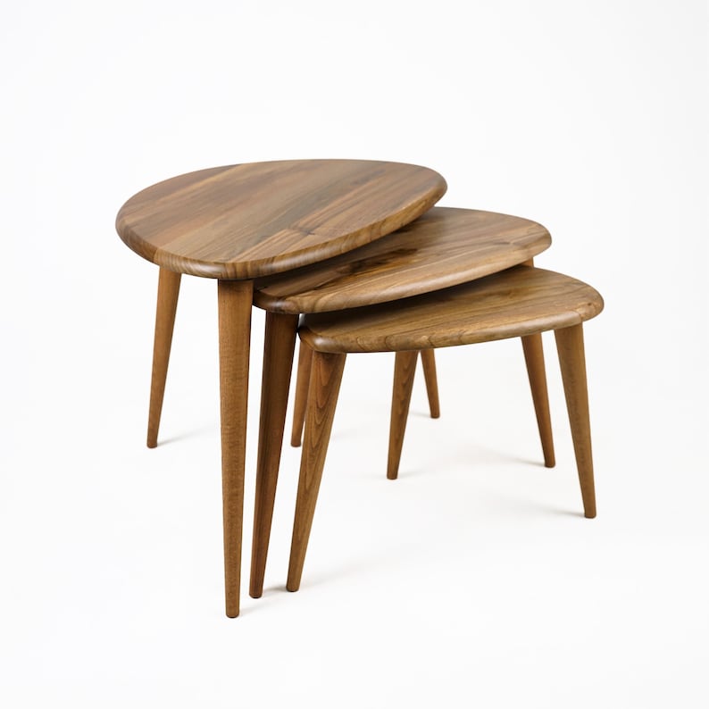 Op de afbeelding: Een set van drie nesttafels gemaakt van hout met een natuurlijke afwerking. De tafels hebben een unieke ovale vorm en worden ondersteund door slanke poten.