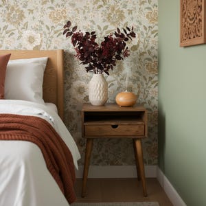 K&ouml;nnte beinhalten: Schlafzimmer-Szene mit einem Nachttisch aus Holz, einer wei&szlig;en, strukturierten Vase mit burgunderfarbenen Zweigen und einem Diffusor f&uuml;r &auml;therische &Ouml;le. Das Bett hat wei&szlig;e Bettw&auml;sche und eine rostfarbene Decke. Blumengemusterte Tapete.