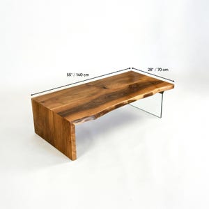 May include: A wooden coffee table with a live edge design. The table is 55 inches long and 28 inches wide. It features a natural wood grain finish and a clear glass leg for support. The table has a modern, rustic aesthetic.