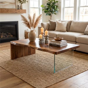 May include: A modern living room featuring a live edge wooden coffee table with a glass leg. The table is on a woven jute rug. Decorative candles and a vase with dried plants are on the table.