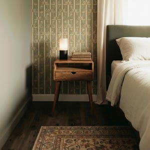 K&ouml;nnte beinhalten: Ein Nachttisch aus Holz im Mid-Century-Modern-Stil mit einer Schublade und einem Regal. Eine kleine Lampe mit schwarzem Sockel und wei&szlig;em Schirm steht oben, zusammen mit einem Stapel B&uuml;cher. Die Szene spielt in einem Schlafzimmer.