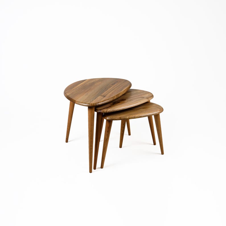 Op de afbeelding: Een set van drie houten nesttafels met een unieke ovale vorm. De tafels zijn gemaakt van natuurlijk hout en hebben een lichtbruine afwerking. De tafels zijn op elkaar gestapeld, met de grootste tafel onderaan en de kleinste tafel bovenaan.