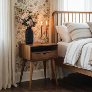 K&ouml;nnte beinhalten: Ein Nachttisch aus Holz mit einer Schublade und einem offenen Regal. Eine schwarze Vase mit Blumen, zwei brennende Kerzen und ein goldener Kerzenhalter stehen darauf. Das Bett hat gestreifte Kissen und eine wei&szlig;e Bettdecke.