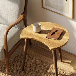 Könnte beinhalten: Kleiner Beistelltisch aus Holz mit halbmondförmiger Platte und drei schrägen Beinen. Eine kleine Keramiktasse und ein braunes, ledergebundenes Notizbuch mit Stift liegen auf dem Tisch.