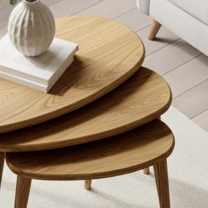 May include: A set of three wooden nesting tables with rounded edges and light brown wood grain. A white vase and a stack of books sit on the top table. The tables are on a white rug.
