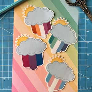 May include: Four rainbow cloud stickers with a sun and a rainbow. The rainbows are in different colours, including purple, blue, green, and orange.