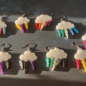 May include: Eight pairs of earrings featuring a white cloud with colourful lightning bolts hanging from it. The earrings are made of resin and have a sparkly finish.