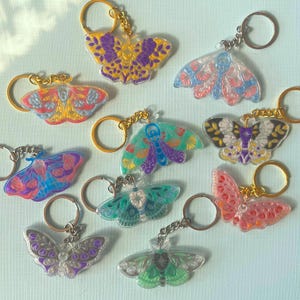 Puede incluir: Un conjunto de diez llaveros de mariposa coloridos. Cada llavero presenta un diseño de mariposa diferente con detalles intrincados y colores vibrantes. Los llaveros están hechos de acrílico transparente y tienen un anillo de metal dorado.