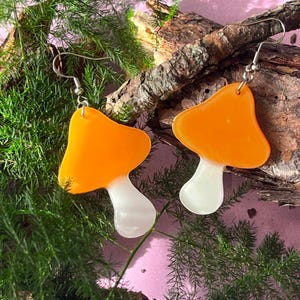 Puede incluir: Un par de pendientes en forma de hongo de color naranja y blanco con ganchos plateados.