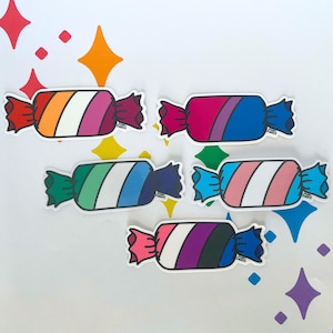 May include: Five candy-shaped stickers, each with a different pride flag design. The stickers have a white central section with coloured stripes, and are wrapped in a colourful wrapper. The background is white with star-shaped decorations.