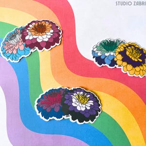May include: A selection of floral stickers in shades of pink, blue, purple, yellow, and green, set against a rainbow-coloured backdrop. The stickers depict detailed illustrations of zinnias with white borders. The text "STUDIO ZABRI" is visible in the top right corner.