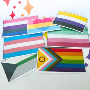 May include: A selection of rectangular stickers showcasing different LGBTQ+ pride flags. The flags feature a variety of colours, such as pink, blue, yellow, purple, and rainbow stripes. Some flags include geometric designs, hearts, and symbols.