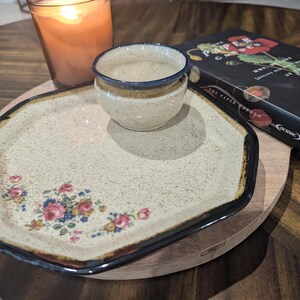 May include: A ceramic plate with a floral design and a small bowl sit on a wooden tray. A lit candle and a book titled "The Paper Garden" are also present. The plate has a speckled beige background with a black border.