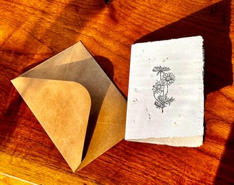 May include: A handmade greeting card featuring a daisy illustration and a brown envelope. The card and envelope are on a wooden surface. The daisy illustration is in black ink. The envelope is open, revealing the card.