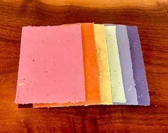 May include: A stack of colorful handmade paper sheets in shades of pink, orange, yellow, green, and purple. The paper has a textured, speckled appearance.