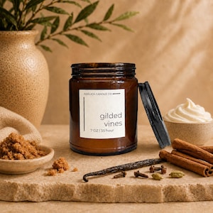 May include: A brown glass candle jar with a white label reading Adelica Candle Co. gilded vines 7 oz | 35 hour, accompanied by a black lid, a small bowl of brown sugar, a vanilla bean, cinnamon sticks, and a small bowl of white cream.