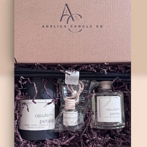 Op de afbeelding: Een geschenkdoos van Adelica Candle Co. met een zwarte kaars, een kleine autodiffuser en een 4 oz geurstokjesdiffuser, allemaal gelabeld Opulent Petals. De items liggen in donkerpaars versnipperd papier in een bruine doos.