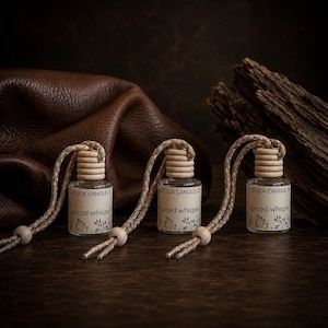 May include: Three small clear glass car air freshener bottles with wooden caps and hanging cords. Each bottle features a beige label with the text Adelica Candle Co and Spiced Whisper, decorated with a subtle botanical leaf illustration.