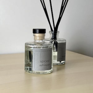 May include: Two clear glass reed diffuser bottles with black reeds. One bottle has a black cap and a grey label that reads "spiced whisper". The other bottle has a black cap and a grey label. The bottles sit on a light wood surface.