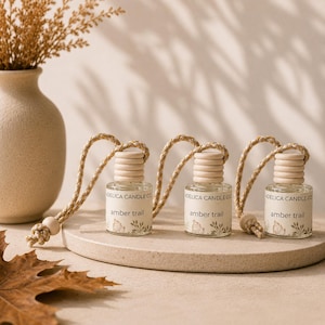 May include: Three small clear glass car air freshener bottles with light wood caps and braided rope hangers. Each bottle features a white label with the text Adelica Candle Co. and amber trail, decorated with small botanical illustrations.