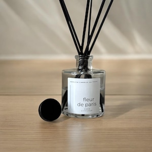 May include: A clear glass reed diffuser with black reeds and a black cap. The label reads "fleur de paris" and "reed diffuser". The diffuser is on a light brown wooden surface.