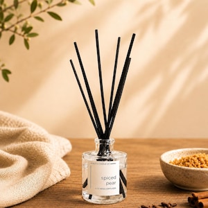 May include: A clear glass reed diffuser bottle with a white label reading Spiced Pear 4 oz Reed Diffuser. The bottle contains a clear liquid and features several thin black reeds extending upwards from the narrow neck of the glass container.
