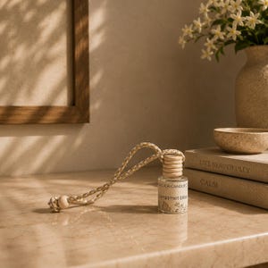 May include: A small glass car air freshener bottle with a light wooden cap and a braided beige hanging cord. The label features the text "ADELICA CANDLE CO" and "bergamot bliss" with a botanical illustration. The bottle is clear and cylindrical.